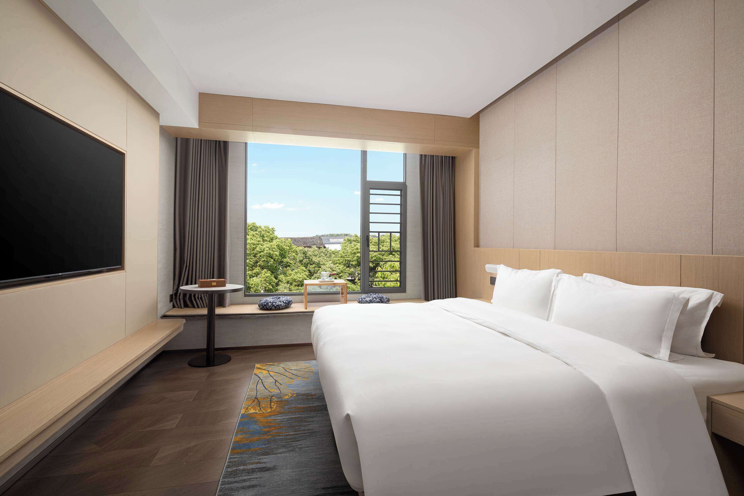 A hotel room with a large bed, a flat-screen TV, a window with curtains, and a view of greenery.