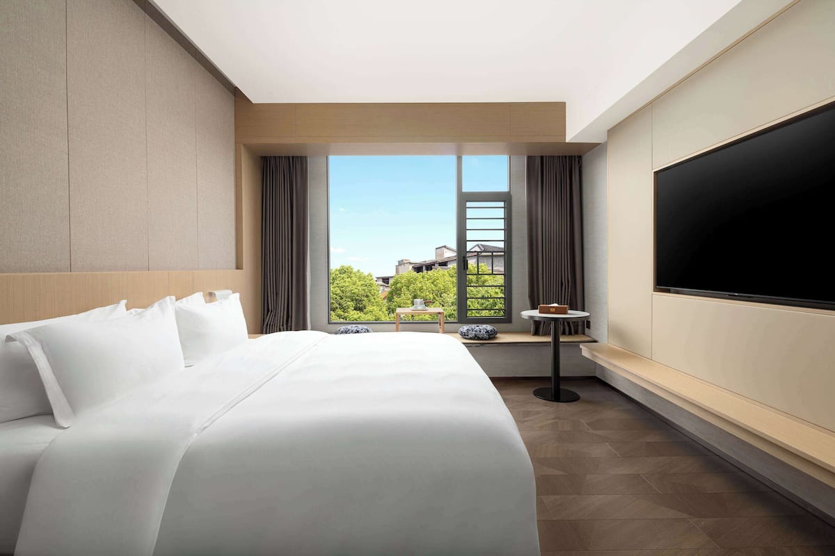 Quarto de hotel com uma cama grande, televisão na parede, uma mesinha e janela com vista para áreas verdes e edifícios.