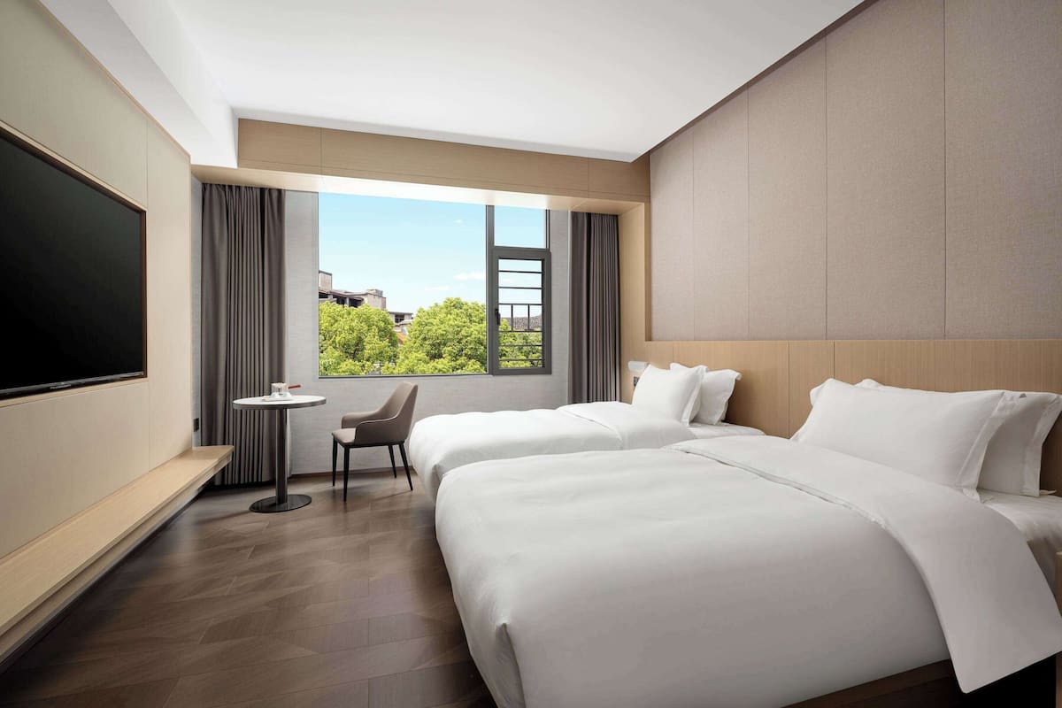 Quarto de hotel com duas camas, televisão, uma mesinha e janela com vista para árvores e edifícios.