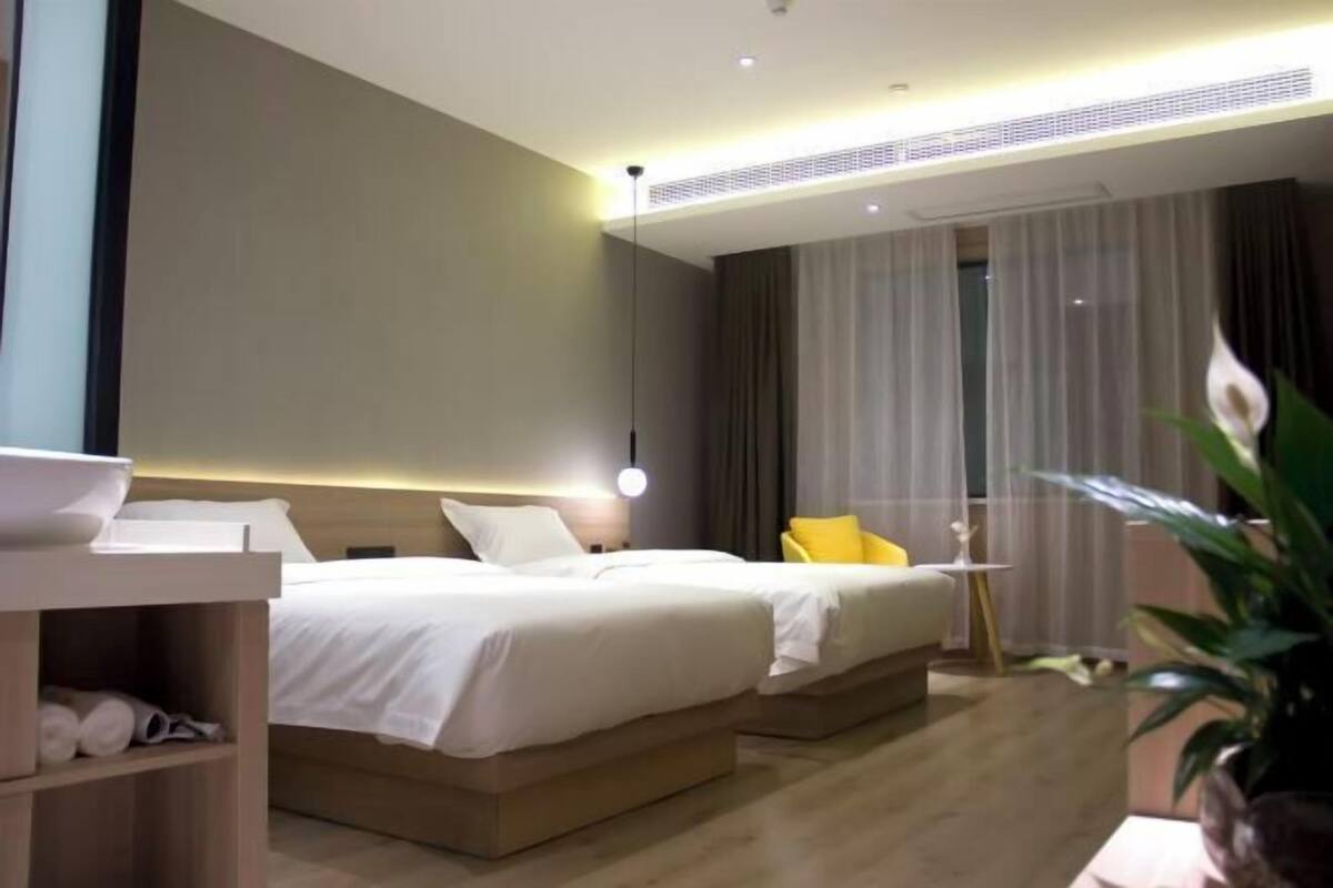 Ein modernes Hotelzimmer mit zwei Betten, einem Nachttisch mit Lampe, einem kleinen Tisch mit einer Vase und einem großen Fenster mit Vorhängen.