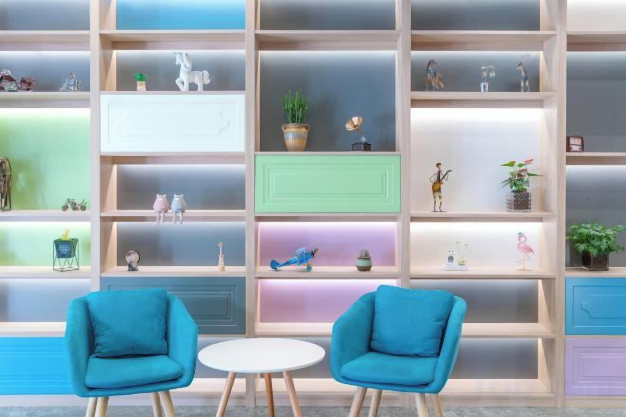 A room with two blue chairs, a white table, and a bookshelf with various items.