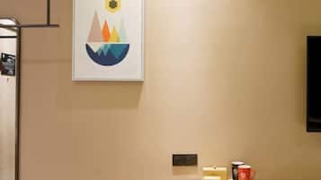 A hotel room with a framed abstract artwork, a small table with a tray, a red mug, and a television mounted on the wall.