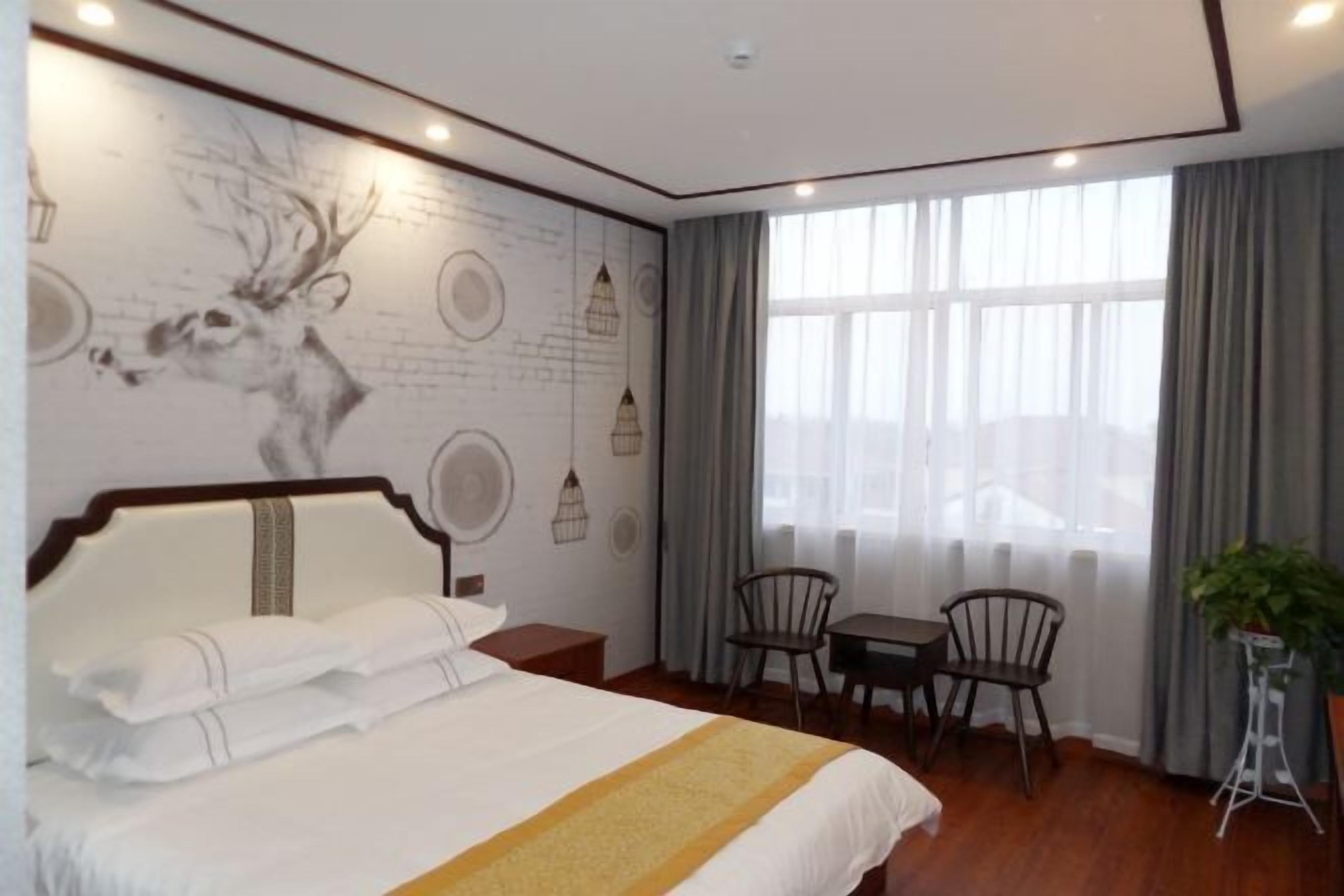 Habitación de hotel con una cama grande, una zona de comedor con dos sillas, una mesita y una planta en maceta, además de un mural en la pared.