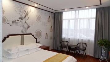 Habitación de hotel con una cama grande, una zona de comedor con dos sillas, una mesita y una planta en maceta, además de un mural en la pared.