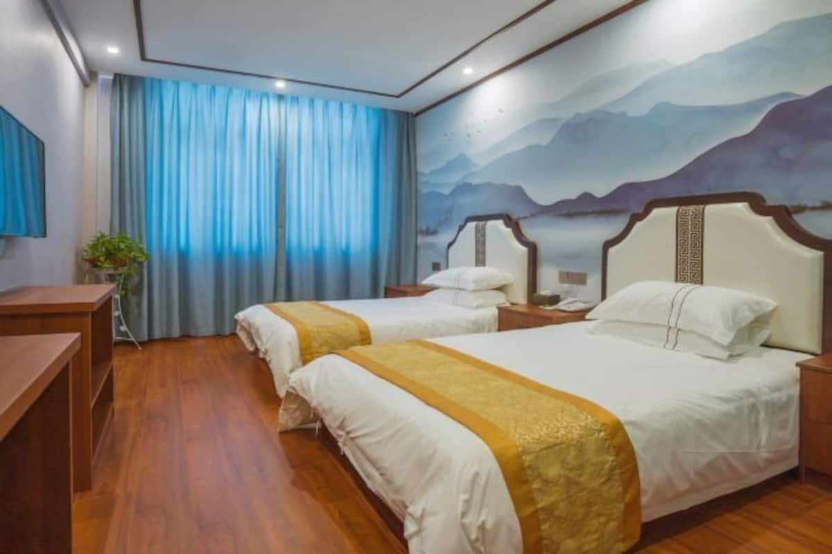 Ein Hotelzimmer mit zwei Betten, Holzboden, einem Schreibtisch und einem Gemälde von Bergen an der Wand.