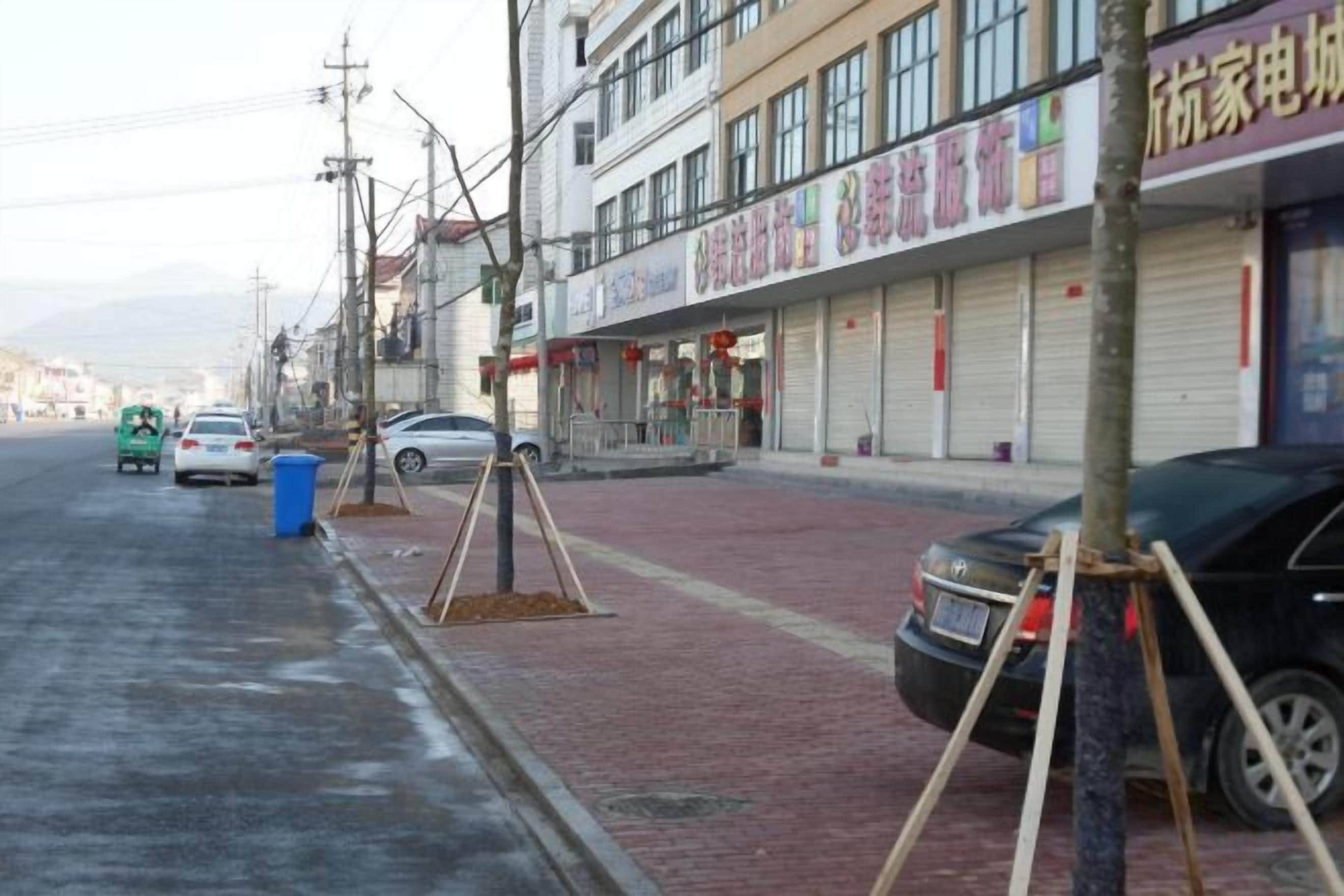 A street with parked cars, a blue trash bin, and a building with closed shutters.