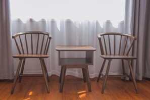 Una habitación con dos sillas de madera y una mesita, una ventana con cortinas translúcidas, y suelo de madera.