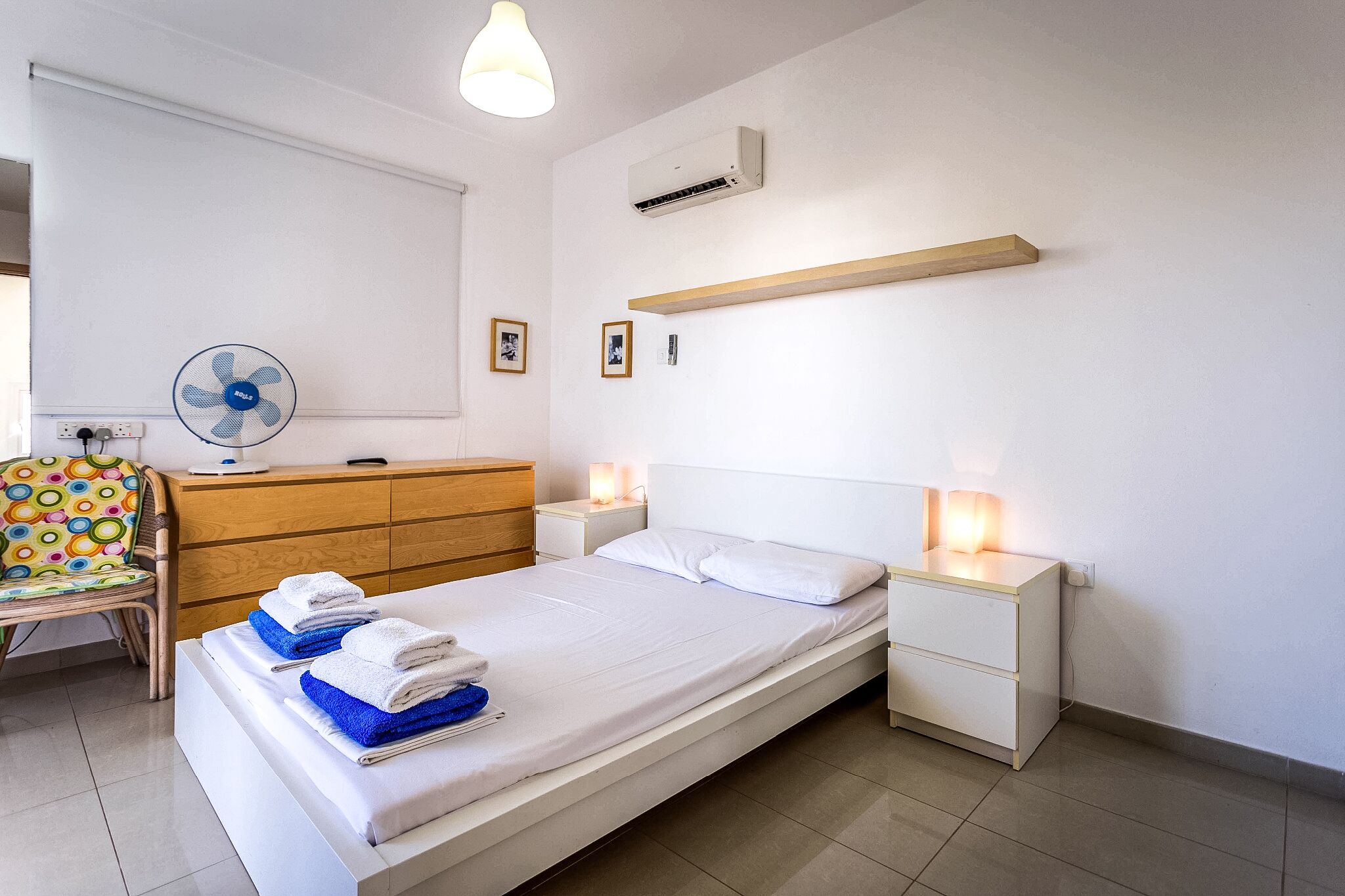 A neatly made bed with white and blue towels, a bedside table with a lamp, a wooden dresser, and a wall-mounted air conditioner.