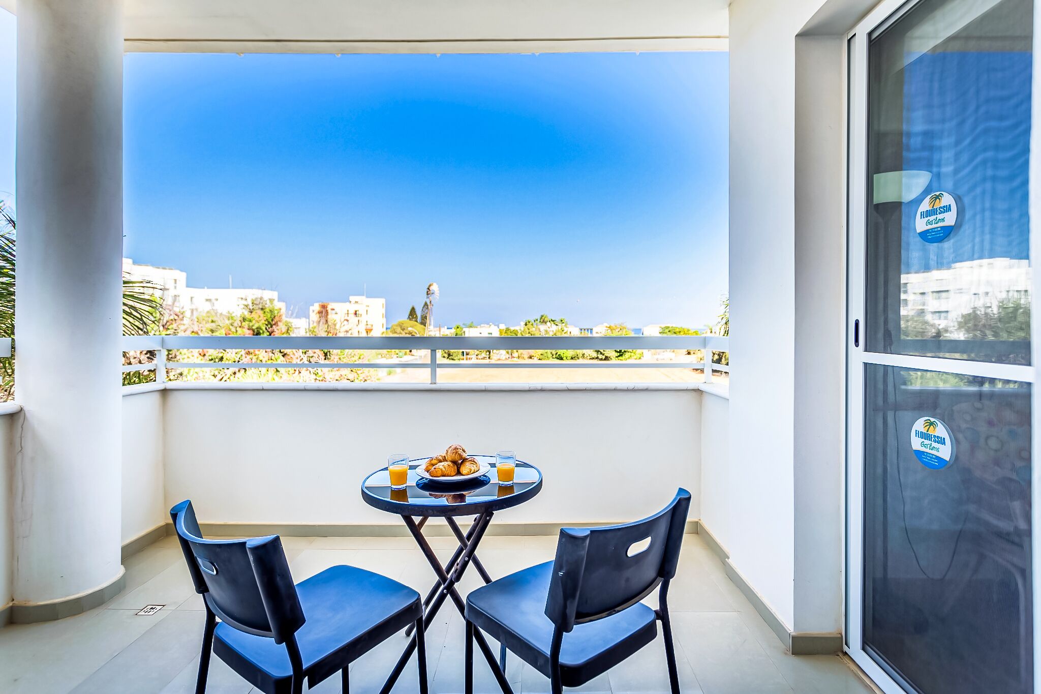 Un balcon avec une table et des chaises, une assiette de pâtisseries et deux verres de jus d’orange.