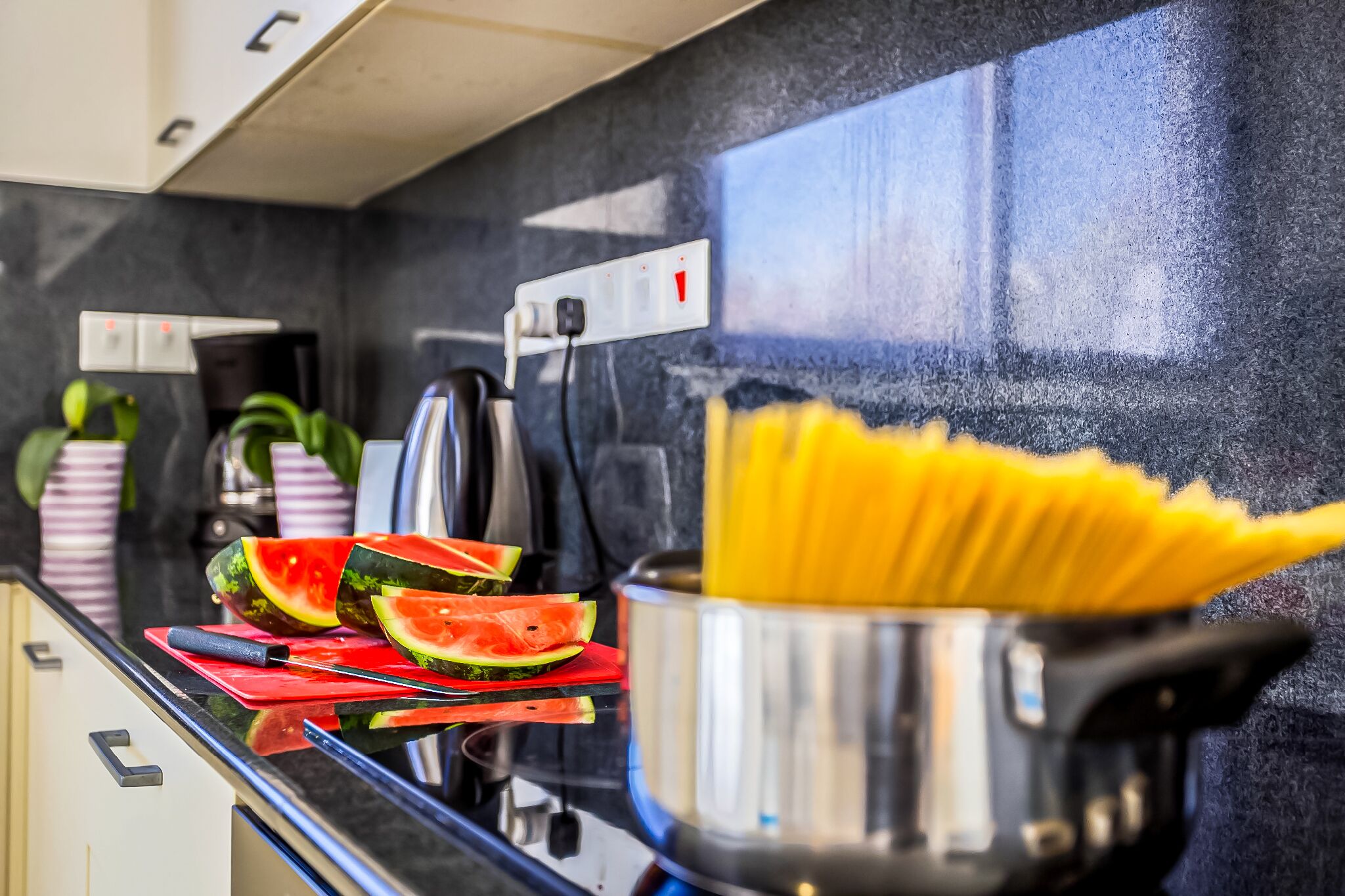 A kitchen counter with a pot of spaghetti, a cutting board with watermelon slices, and a knife.