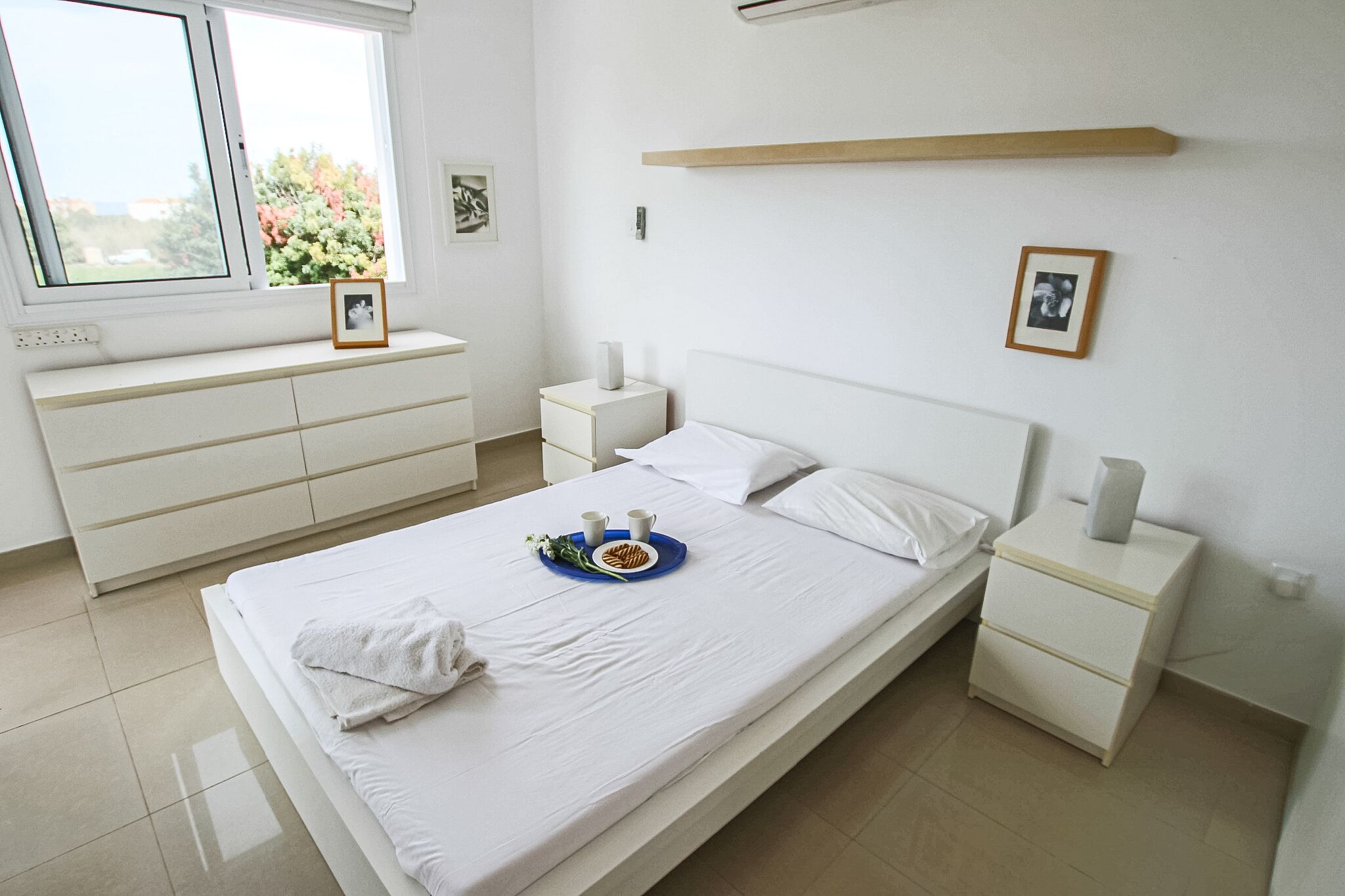 Ein sauberes, modernes Schlafzimmer mit einem Bett, Nachttischen, einer Kommode und einem Fenster mit Blick auf Bäume.