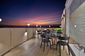A rooftop terrace with wooden flooring, round tables, and black chairs, featuring a sunset view. - HOTEL SURAT (Surat)