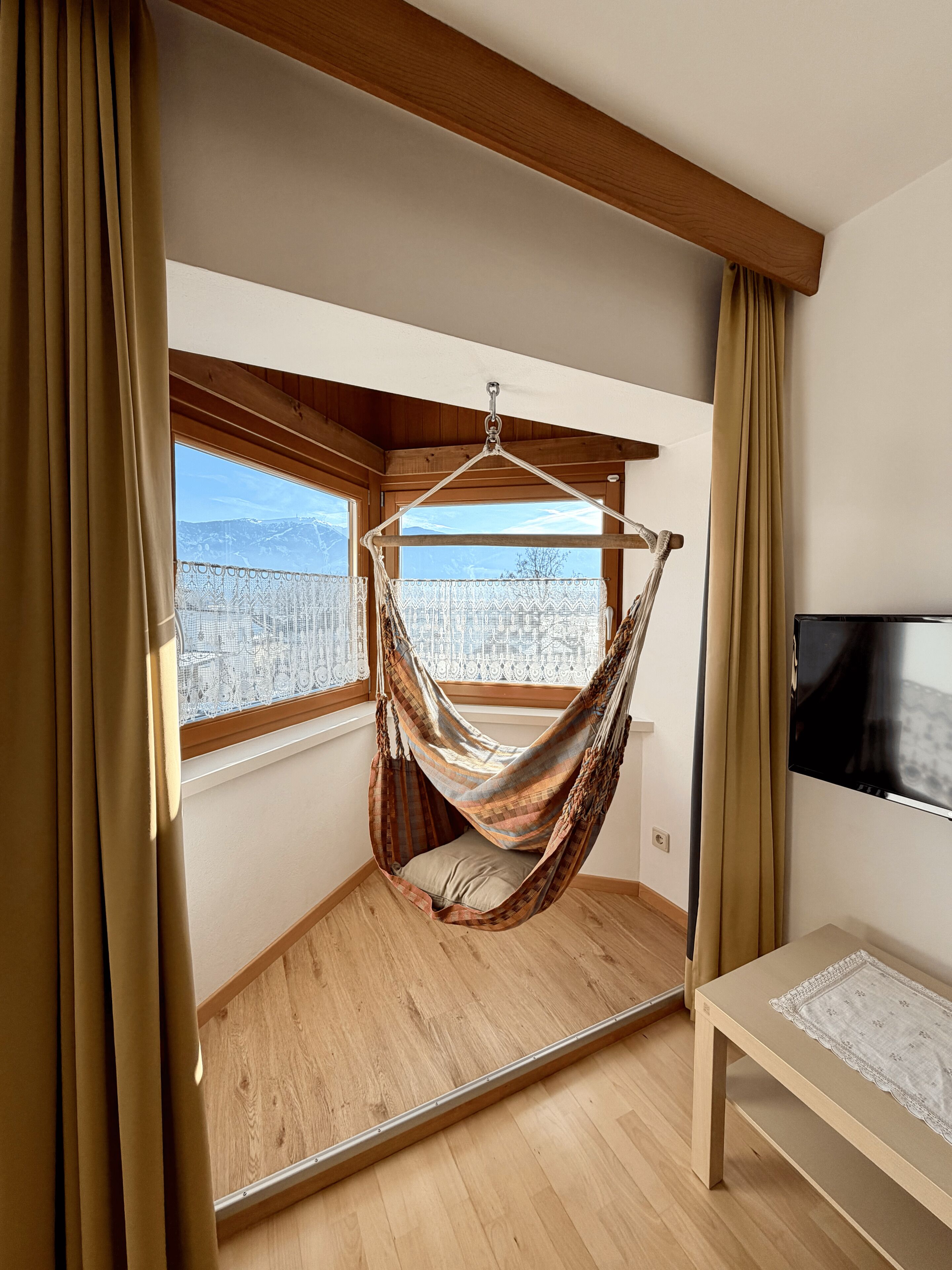 A room with a hammock chair, a wooden floor, a TV, and a window with a view of mountains.