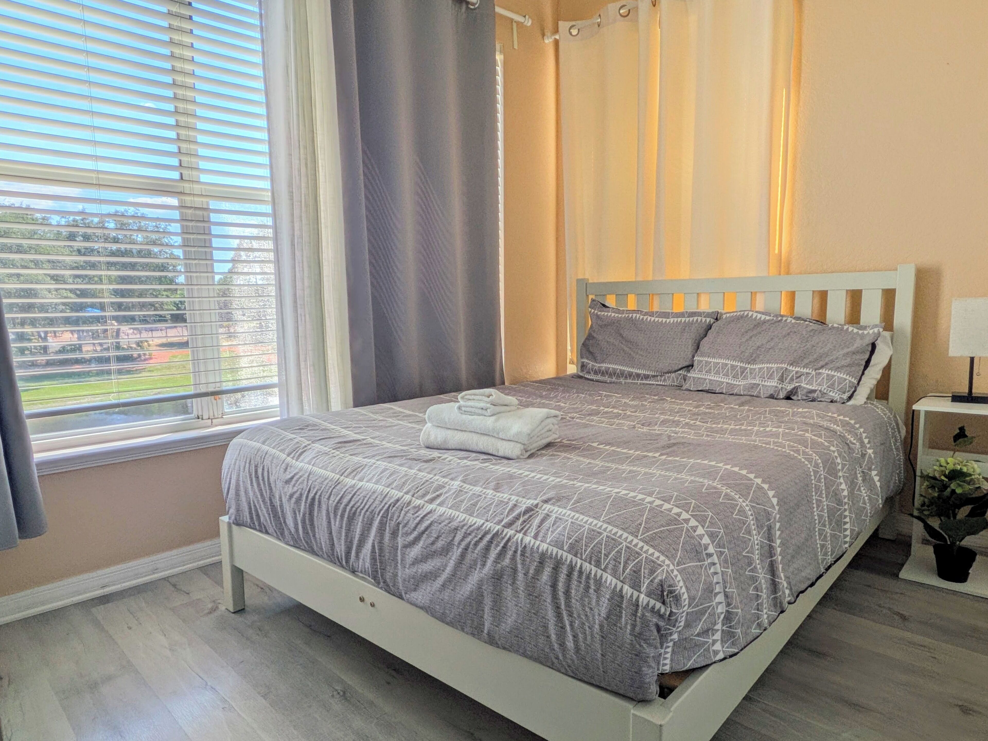 Un dormitorio con cama, cortinas, una ventana con persianas, una mesita de noche con lámpara y una planta en maceta.