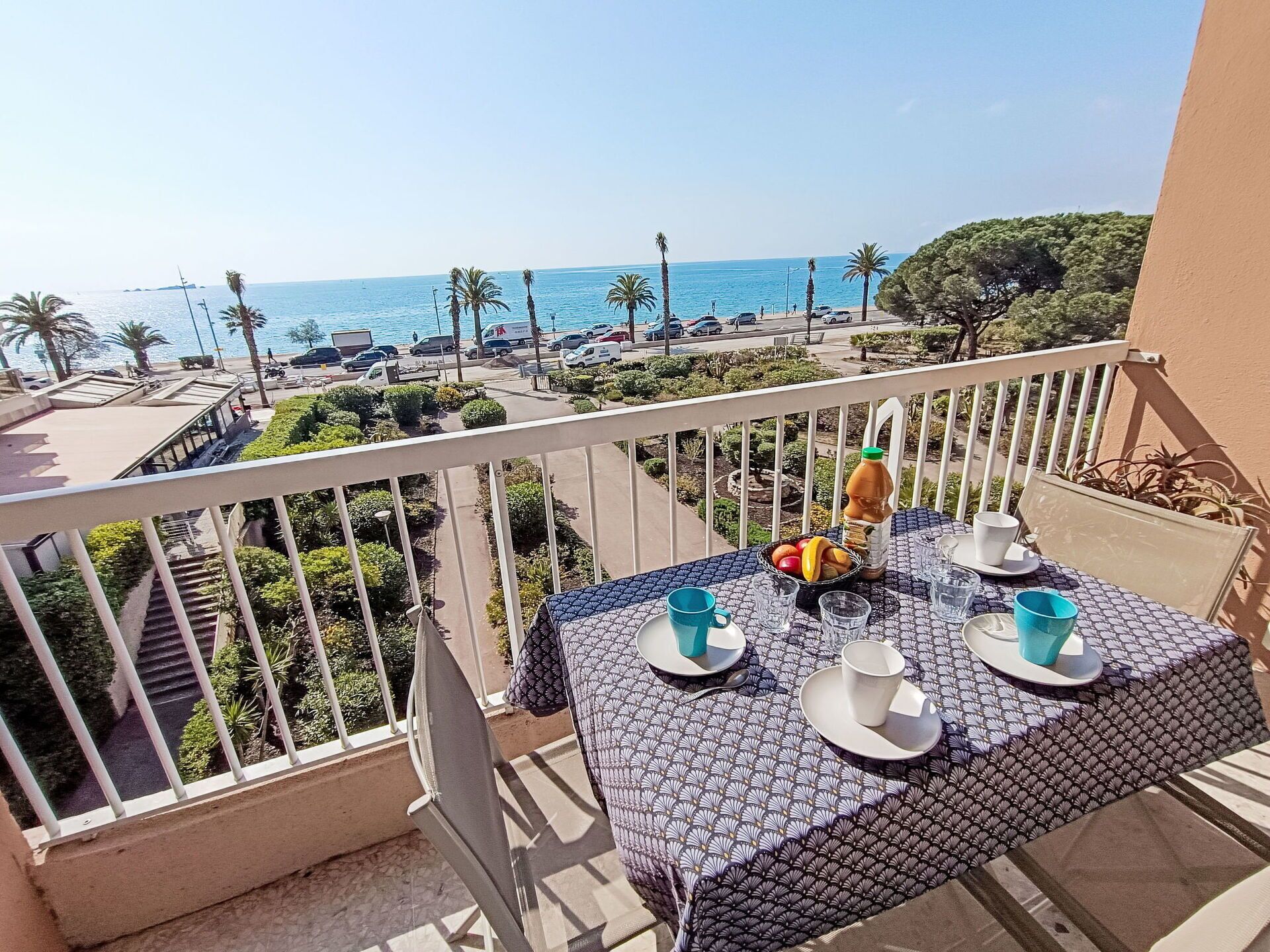 A balcony with a table set for two, overlooking a coastal road with parked cars and palm trees.