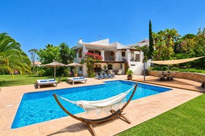 Un espace piscine avec un hamac, des chaises longues et un bâtiment blanc doté d’un toit en tuiles.