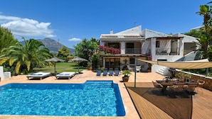 Una piscina con sillas de descanso, una terraza de madera y una casa con fachada blanca y techo de tejas.