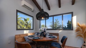 Un espace repas avec une table ronde en bois, dressée pour quatre personnes, entourée de chaises, d’un banc et d’un luminaire suspendu.