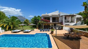 Une piscine avec des chaises longues, une terrasse en bois et une maison à la façade blanche et au toit recouvert de tuiles.