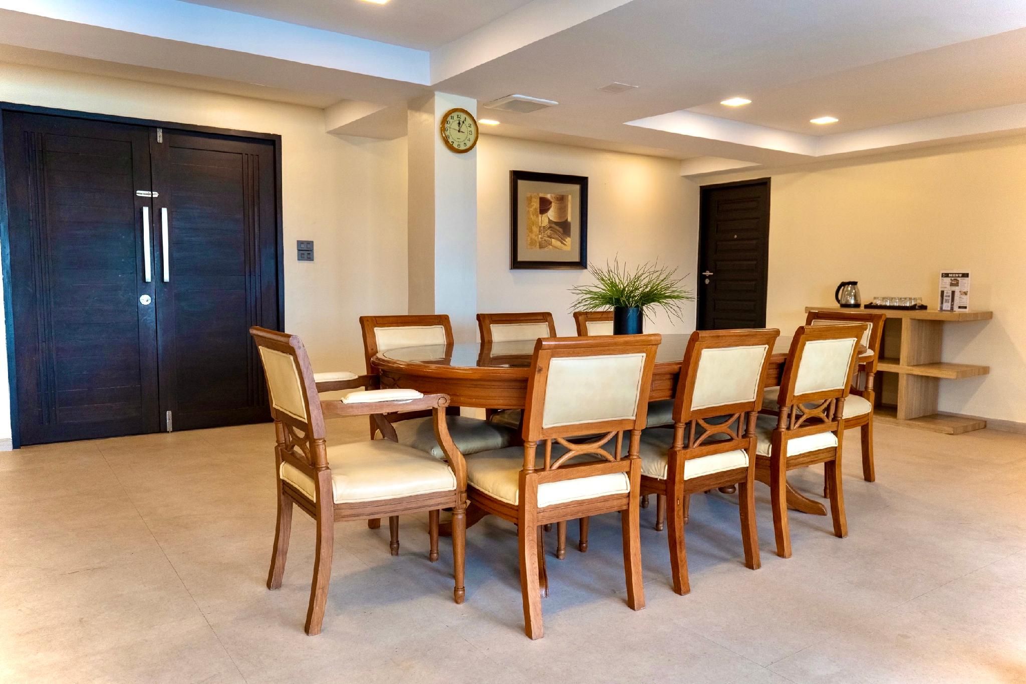 Un coin repas avec une table ronde entourée de chaises en bois dotées d’assises rembourrées, une horloge au mur et une porte à cadre sombre.