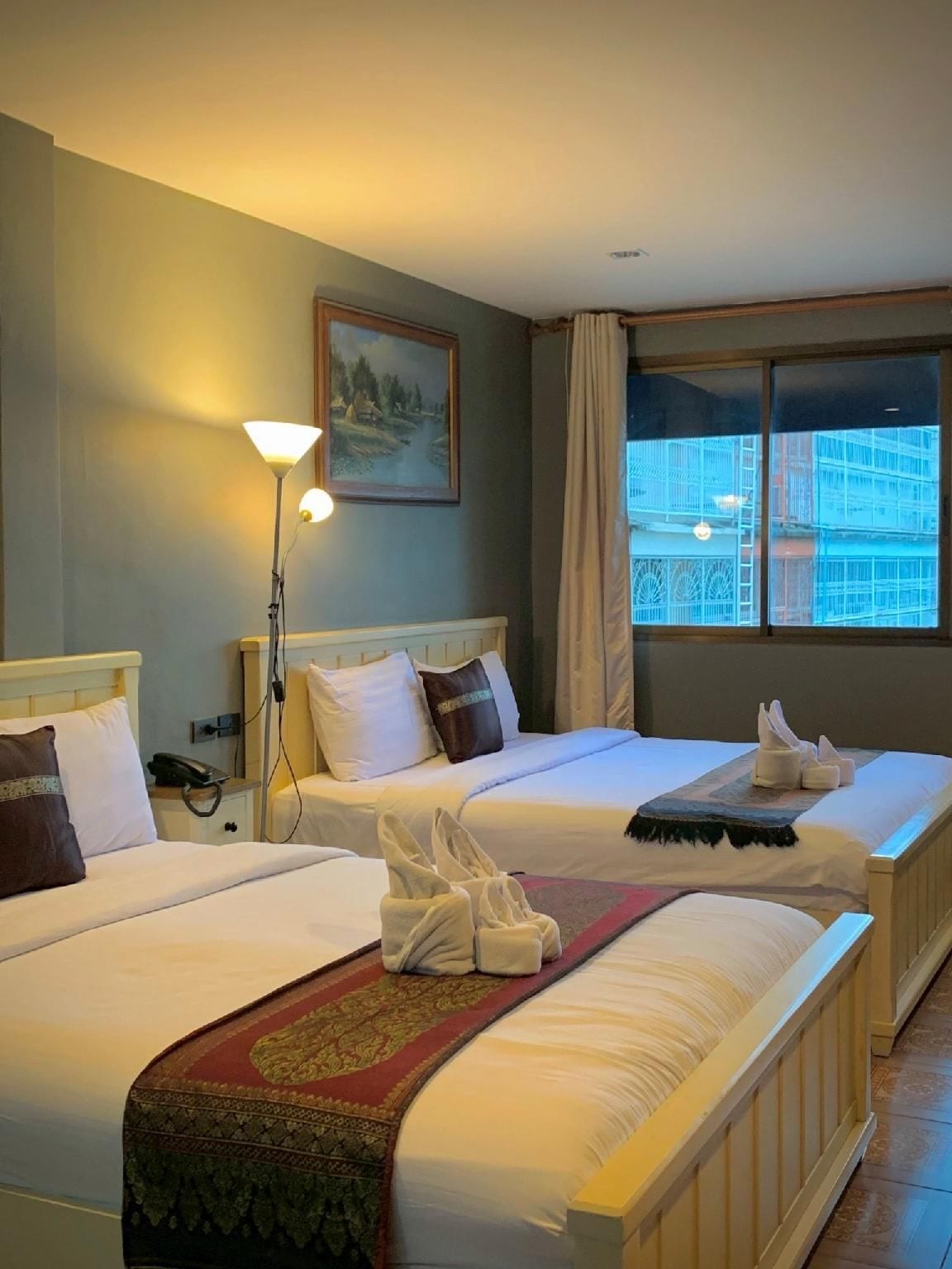A hotel room with two beds, a painting on the wall, a lamp, and a window with a view of a building.