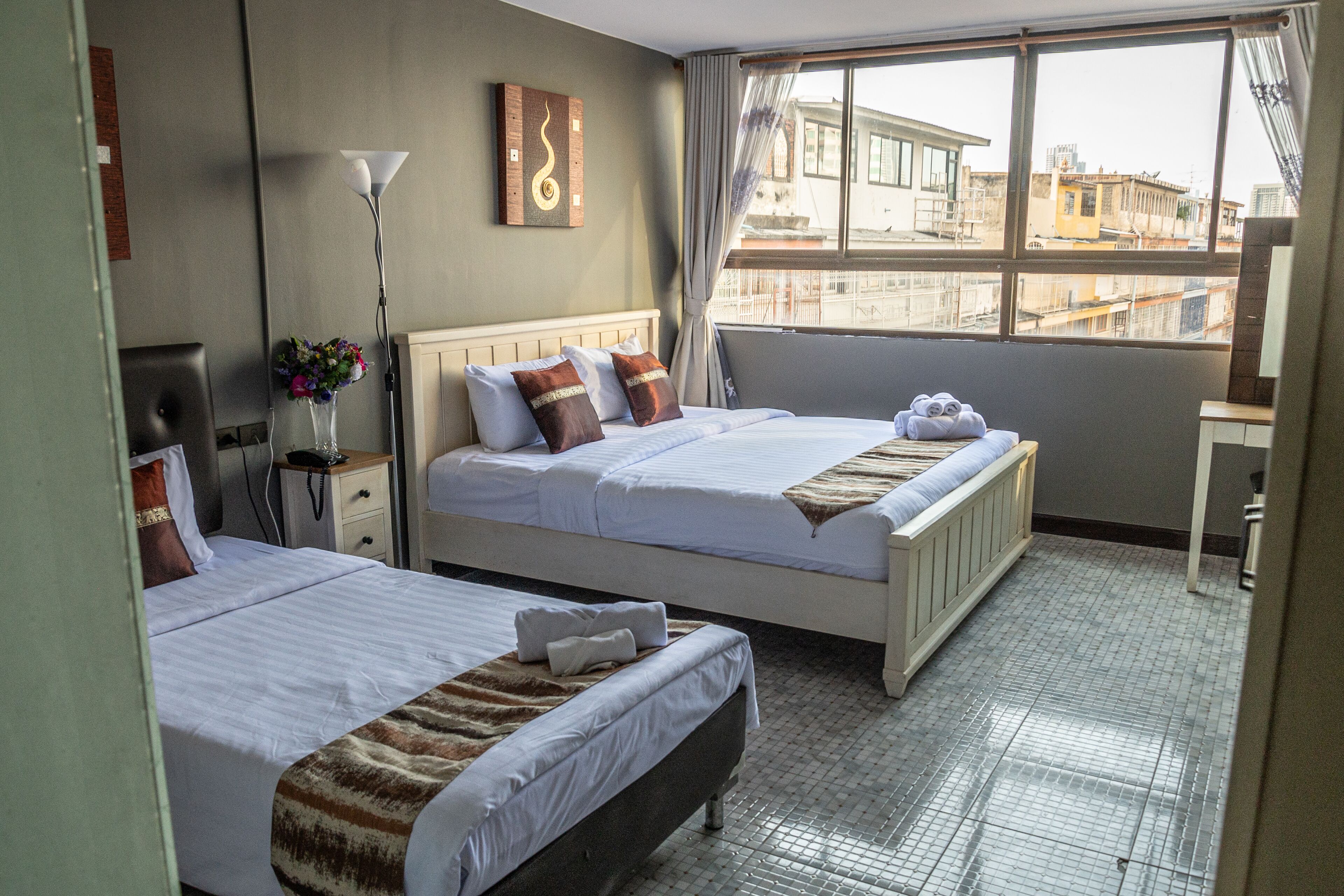 A hotel room with two beds, a large window with a view of buildings, a floor lamp, and a wall art piece.