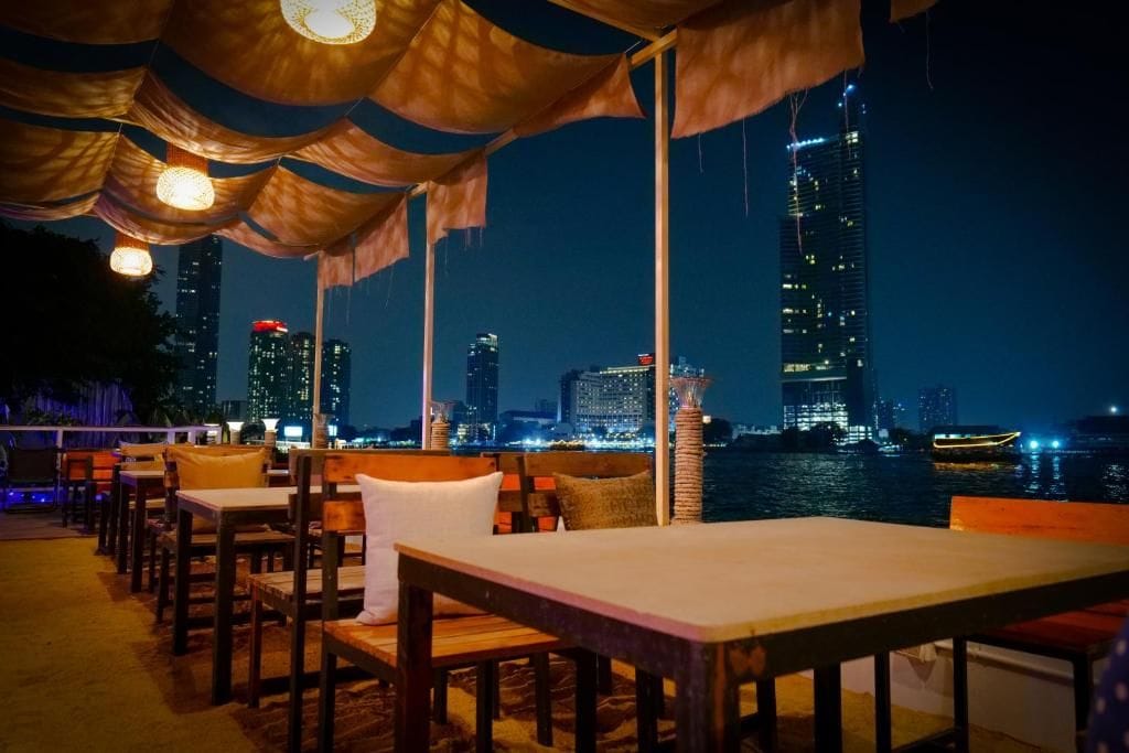 Un área de comedor al aire libre con mesas y sillas, con vistas a un horizonte urbano nocturno.
