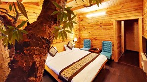 A wooden cabin room with a bed, two blue chairs, and a tree in the corner. - Treehouse Resort (Dhari)