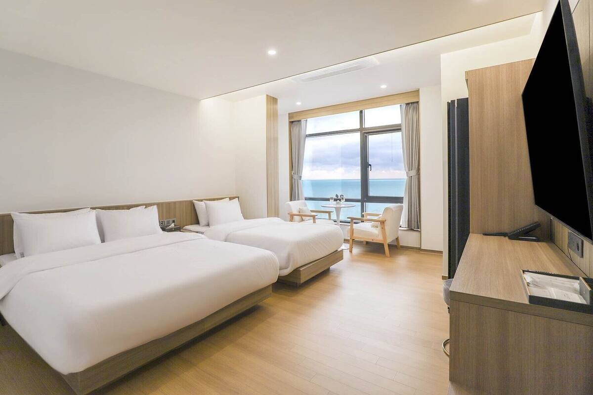 Habitació d'hotel amb dos llits, un escriptori, una cadira, un televisor i un gran finestral amb vista al mar.