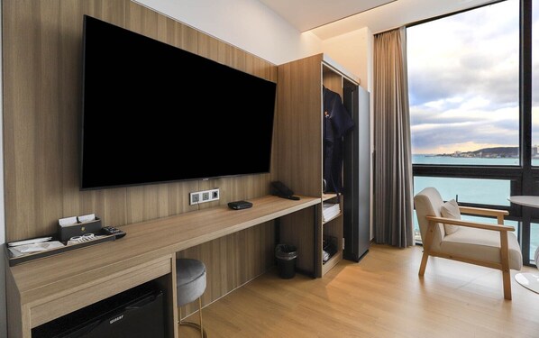Une chambre d’hôtel moderne dotée d’un grand téléviseur à écran plat fixé au mur, d’un bureau en bois avec un ordinateur portable, d’une chaise et d’une vue sur la mer par une grande fenêtre.