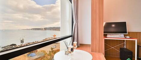 A modern hotel room with a large window offering a view of the sea and a distant cityscape. There's a round white table with a glass of water and a bottle, a TV on a stand, and a computer on a desk.