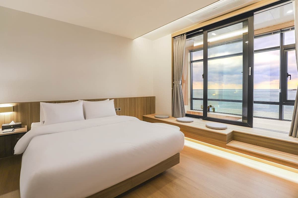 Habitació d'hotel amb un llit gran, panell de capçal de fusta, taules de nit i vista a l'oceà a través d'una finestra ampla.