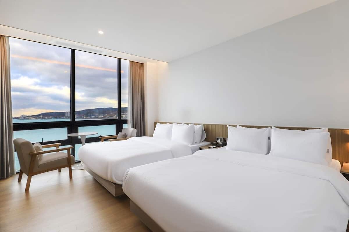 Habitació d'hotel amb dos llits, una cadira, una taula petita i vista al mar.