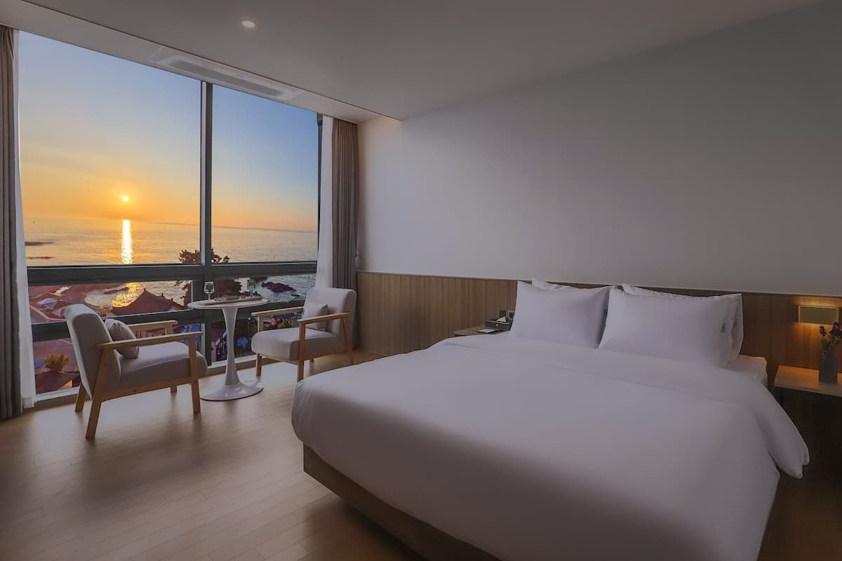 Habitació d'hotel amb un llit gran, un escriptori amb cadires i vista cap al mar mentre se pon el sol.