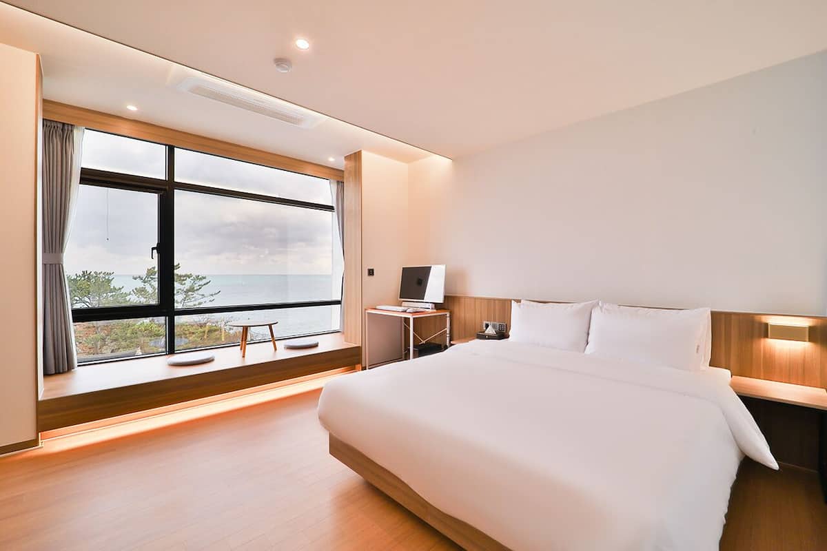 Habitació d'hotel moderna amb un llit gran, una taula de nit, un escriptori i vista al mar a través d'una finestra ampla.