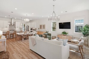 Amplia sala de estar con sofá blanco, mesa y sillas de comedor, cocina con armarios blancos y un ventanal grande.