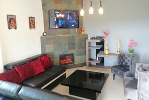 Uma sala de estar com um sofá preto, almofadas estampadas vermelhas, uma mesa de centro preta, uma lareira, uma TV fixada na parede e vasos decorativos em uma estante.