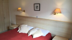 Una cama bien tendida con colcha roja y almohadas blancas, dos lámparas de noche, un cuadro enmarcado en la pared y un armario de madera.