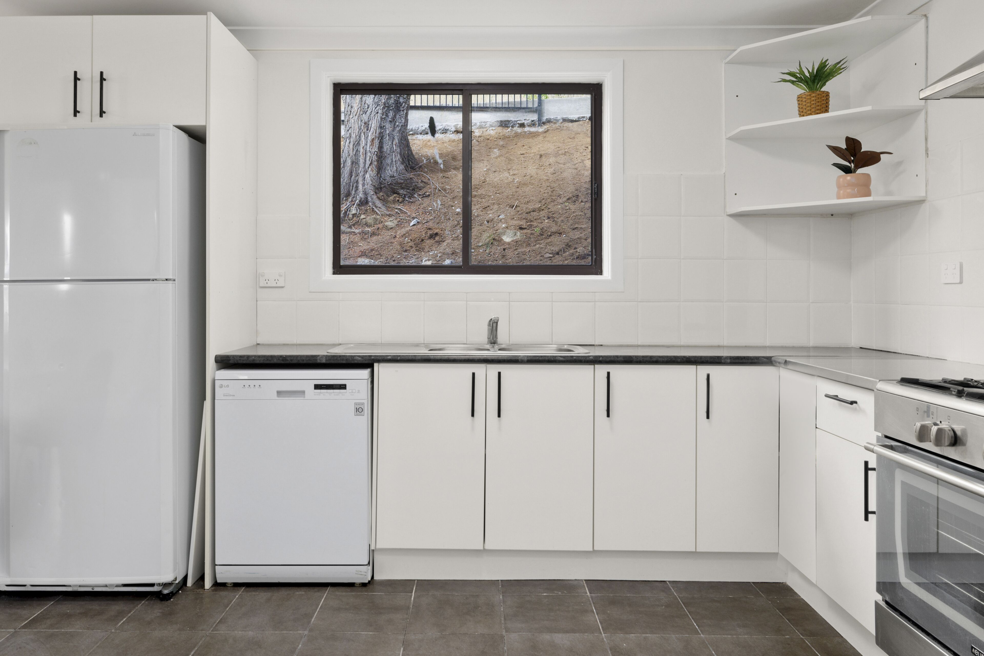 A modern kitchen with white cabinets, a dishwasher, a stove, and a window with a view of a tree and fence.