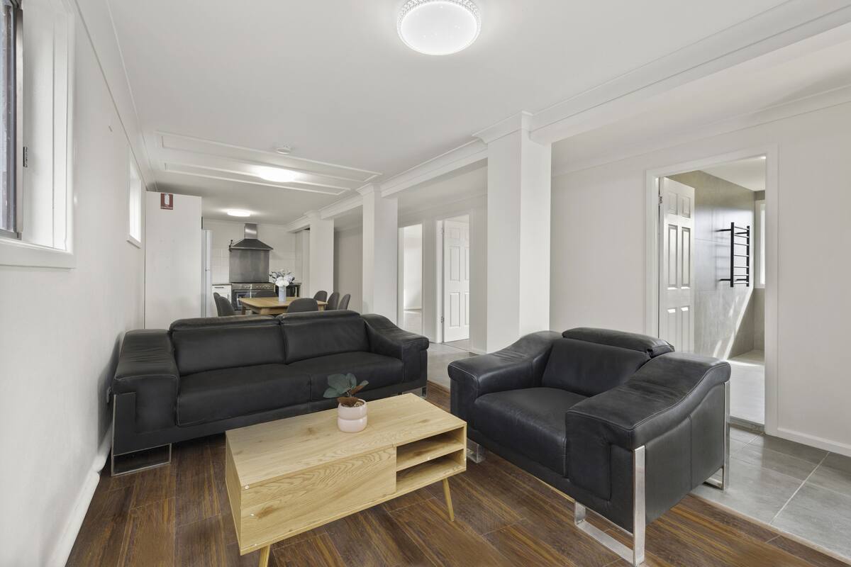 A modern living room with a black leather sofa, a wooden coffee table, and a black armchair. A kitchen area is visible in the background.