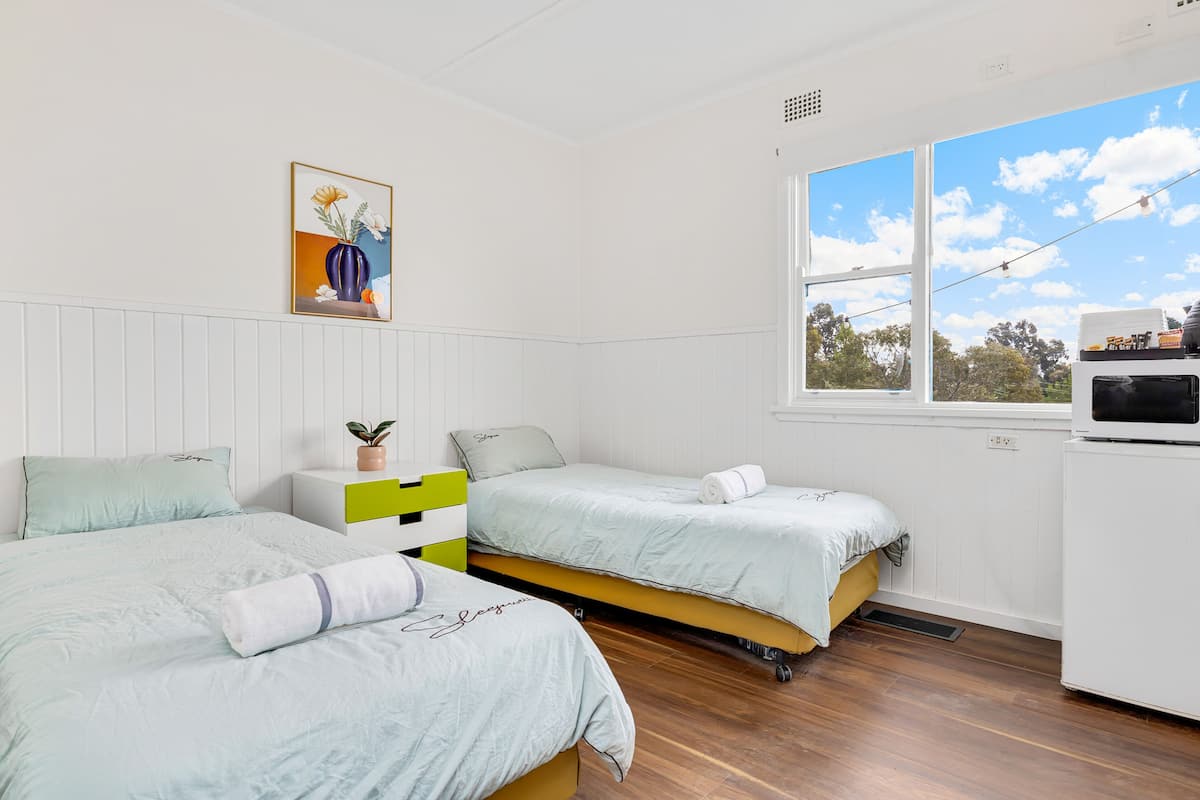 A bedroom with two beds, a window with a view, a painting on the wall, and a white appliance.