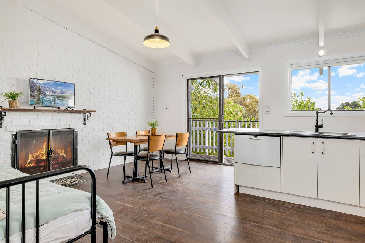 A modern kitchen with white cabinets, a dining area with wooden chairs, a fireplace, and a large window with a view of greenery.