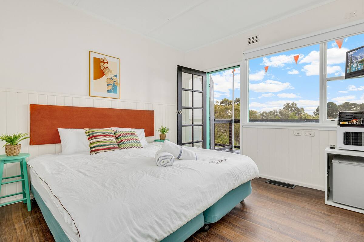 A bedroom with a large bed, a wooden headboard, a small table with a plant, and a window with a view of the outdoors.