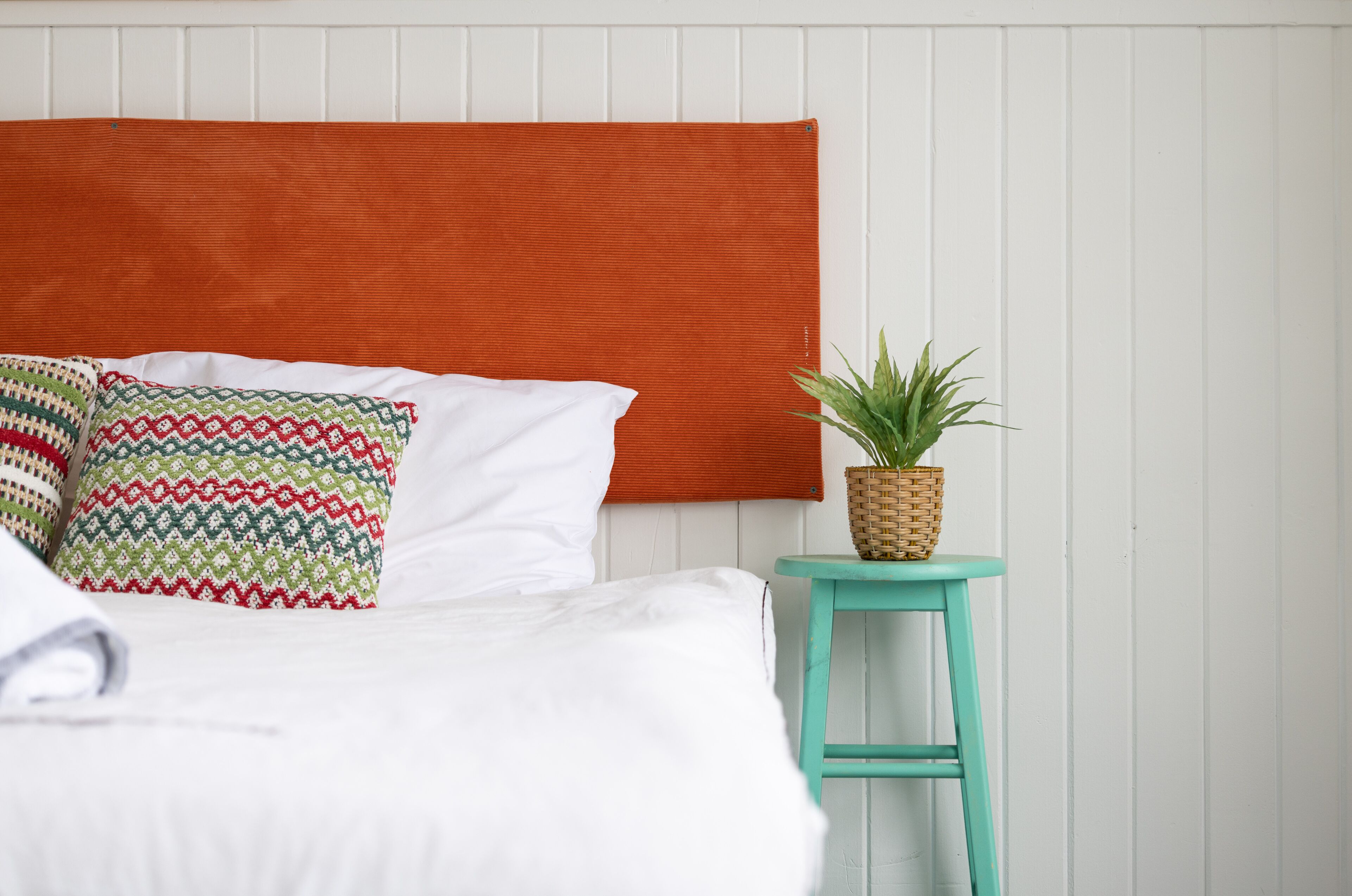 A bed with white bedding and patterned pillows, an orange headboard, and a small teal stool with a plant.