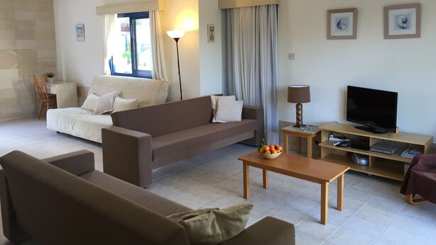 A modern living room with a sofa, a coffee table, a TV, and a lamp. - One Bedroom Bungalow Deluxe - Flouressia Gardens (Protaras)