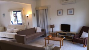 A living room with a sofa, armchair, coffee table, TV stand, and a bowl of fruit. - One Bedroom Bungalow Deluxe - Flouressia Gardens (Protaras)