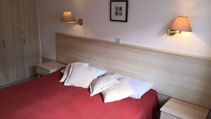 A neatly made bed with a red bedspread and white pillows, two bedside tables with lamps, a wooden headboard, and a framed picture on the wall. - One Bedroom Bungalow Deluxe - Flouressia Gardens (Protaras)