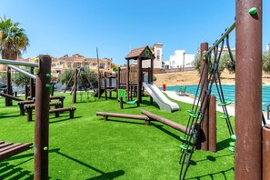 A playground with various play structures, including a slide and climbing elements, set on a green artificial turf.