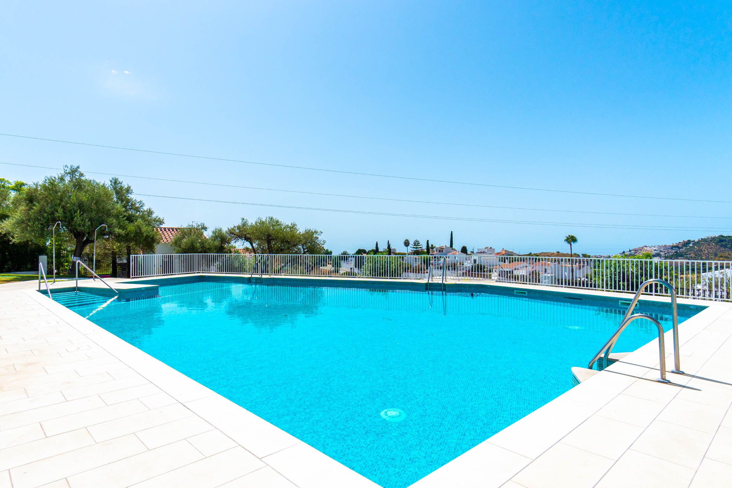 A rectangular swimming pool with a white tiled deck, surrounded by a white fence, and a clear blue sky above.