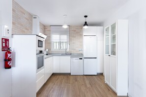 A modern kitchen with white cabinetry, a built-in oven, a microwave, a dishwasher, and a refrigerator. There is a fire extinguisher mounted on the wall, and a window with a view of the outside.